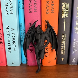 May include: A black figurine of a winged male figure stands in front of a row of colorful books. The figurine has detailed wings and is posed with one leg slightly bent. The books have titles and author names printed on their spines.