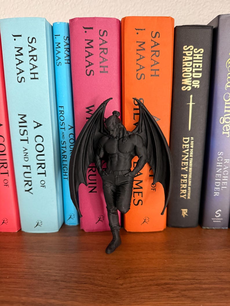 May include: A black figurine of a winged male figure stands in front of a row of colorful books. The figurine has detailed wings and is posed with one leg slightly bent. The books have titles and author names printed on their spines.