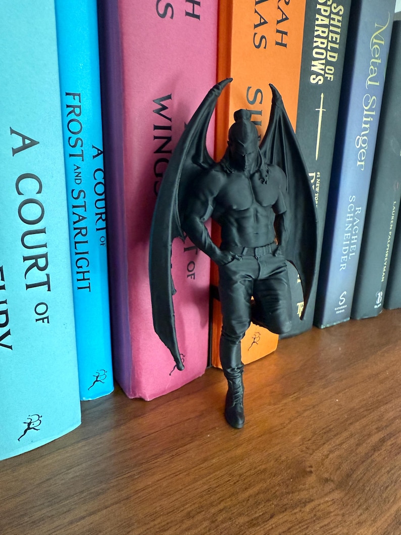 May include: A black figurine of a winged humanoid figure, leaning against a row of colorful books. The figure has a muscular physique, a helmet, and large, spread wings. The books have titles like "A Court of Frost and Starlight" and "Metal Slinger."