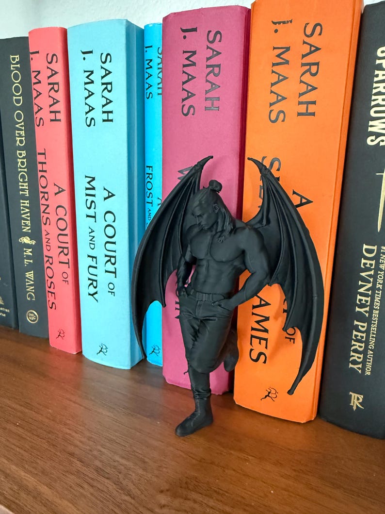 May include: A black figurine of a muscular winged figure leans against a shelf of colorful books. The books have titles like "A Court of Thorns and Roses" and "A Court of Mist and Fury" by Sarah J. Maas. The figurine has detailed wings.