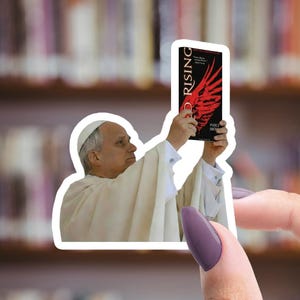 May include: A sticker featuring a figure in white robes holding up a book titled "Red Rising" with a red wing design. The background shows a blurred library setting.
