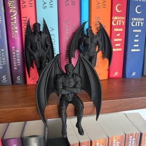 May include: Three matte black figurines of winged humanoid characters. Two figures stand upright with wings spread, while one figure sits with arms crossed and wings extended. Each character features detailed musculature and clothing textures.