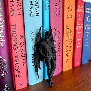 Puede incluir: Una figura negra mate de un personaje masculino alado de pie con las manos en los bolsillos, colocada entre una fila de libros coloridos. Los libros incluyen t&iacute;tulos de Sarah J. Maas como A Court of Thorns and Roses, Mist and Fury y Silver Flames.