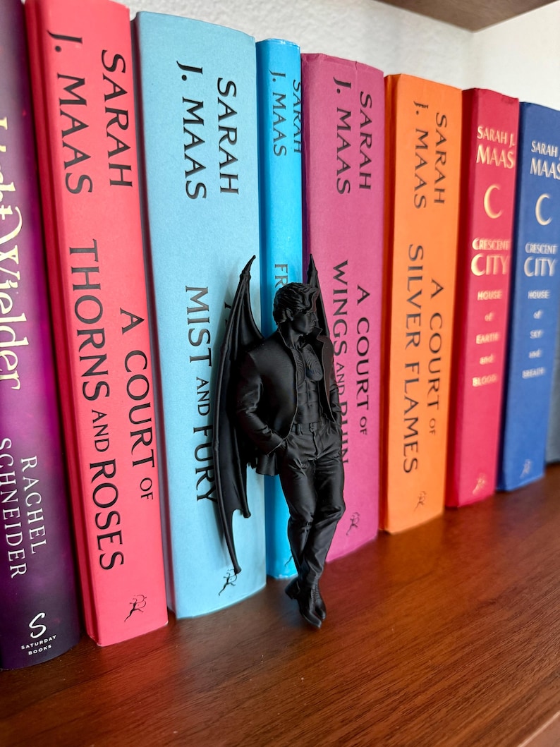 Puede incluir: Una figura negra mate de un personaje masculino alado de pie con las manos en los bolsillos, colocada entre una fila de libros coloridos. Los libros incluyen t&iacute;tulos de Sarah J. Maas como A Court of Thorns and Roses, Mist and Fury y Silver Flames.