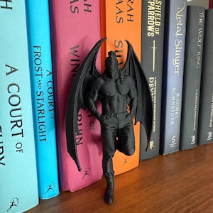 May include: A black figurine of a muscular winged figure with a helmet, standing in front of a row of colorful books. The figurine is detailed with visible muscles and wings. The books have titles like "A Court of Frost and Starlight" and "Shield of Sparrows."