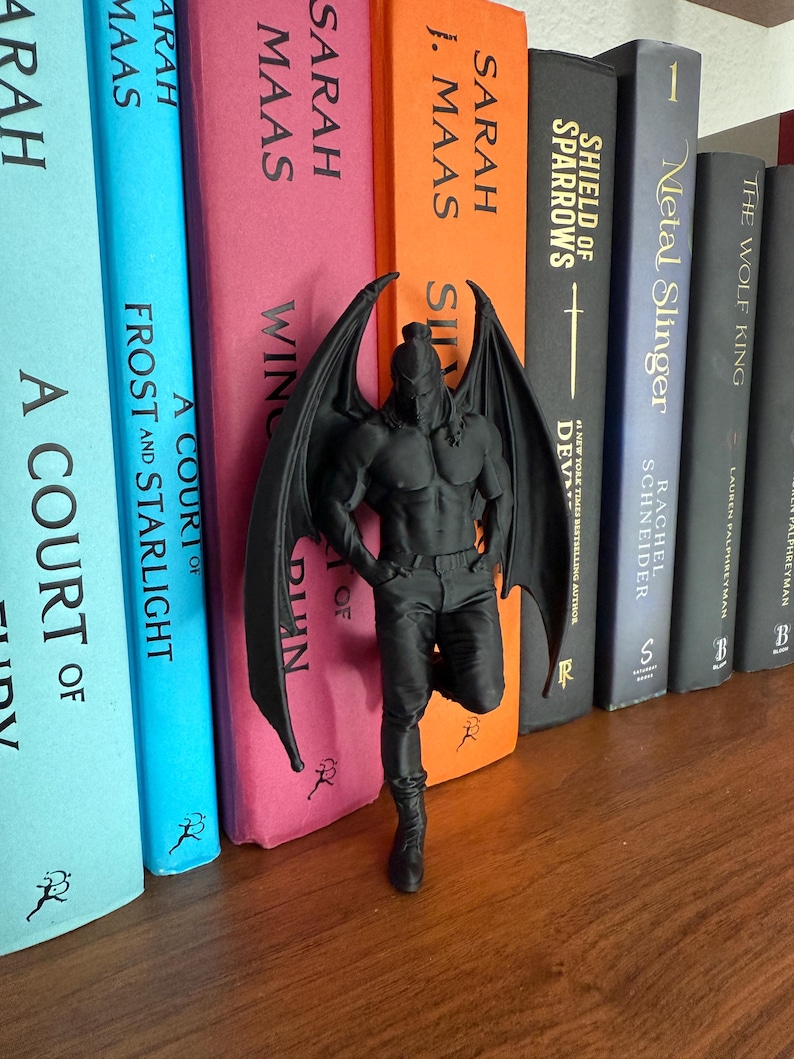 May include: A black figurine of a muscular winged figure with a helmet, standing in front of a row of colorful books. The figurine is detailed with visible muscles and wings. The books have titles like "A Court of Frost and Starlight" and "Shield of Sparrows."