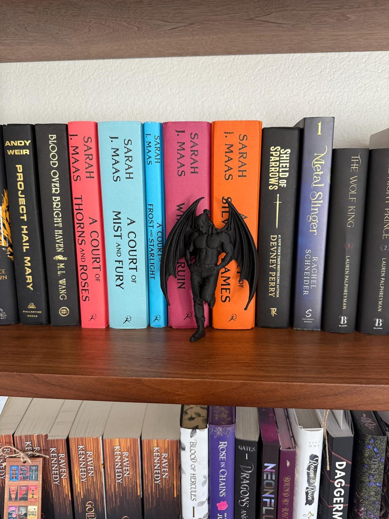 May include: A bookshelf displays a collection of books with colorful spines. A black figurine of a winged creature stands in front of the books. The books are arranged on two wooden shelves, with various titles and authors visible. The overall theme is fantasy literature.
