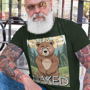 May include: Dark green t-shirt featuring a cartoon bear with the words "BEAR NAKED" above and below the bear. The bear has a sad expression and is standing in front of a forest scene. The shirt is short-sleeved.
