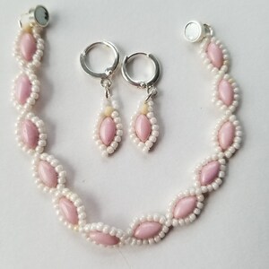 May include: A handmade jewelry set featuring a bracelet and earrings. The bracelet has a magnetic clasp and is made with pink oval beads and white seed beads. The earrings have a teardrop design with matching pink and white beads. The set is on a white background.