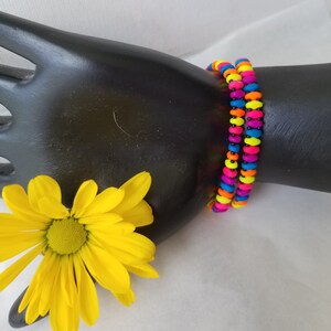 May include: Two colorful bracelets made of neon pink, yellow, blue, and black beads. The bracelets are stacked on top of each other on a black mannequin arm.