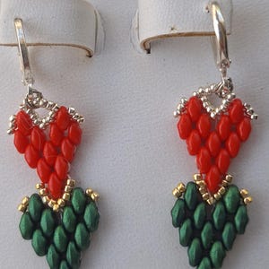 May include: A pair of handmade earrings featuring a red heart-shaped top and a green leaf-shaped bottom. The earrings are made with small beads and silver-colored metal hooks. The design is detailed and the colors are vibrant.