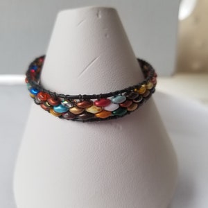May include: A multi-colored beaded bracelet with a black wire base. The beads are small and round, and they are arranged in a pattern. The bracelet is on a white display stand.