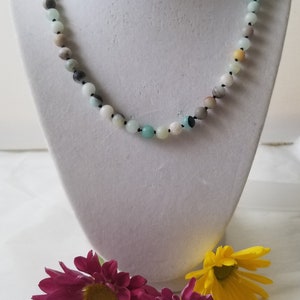 May include: A necklace made of green, blue, and brown beads on a black string. The necklace is displayed on a white mannequin bust. Three pink and yellow flowers are in the foreground.