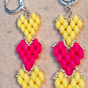 May include: A pair of handmade earrings featuring a heart design. The earrings are composed of yellow and pink beads, with a silver-colored clasp. The heart shapes are made of small, oval-shaped beads.