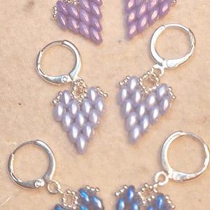 May include: Three pairs of heart-shaped earrings in shades of purple and blue. Each earring features a cluster of teardrop-shaped beads, suspended from silver-colored leverback hoops. The earrings are displayed on a neutral-toned surface.