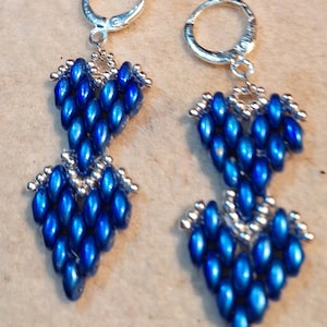 May include: A pair of dangle earrings featuring a heart-shaped design. The earrings are made of small, shiny, blue beads and silver-colored metal accents. The earrings have a lever-back closure.