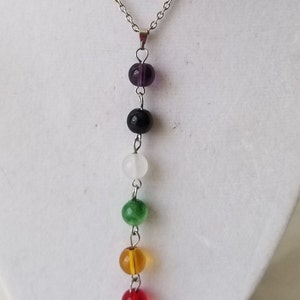 May include: A silver chain necklace featuring seven colorful gemstone beads in a vertical row. The beads are purple, black, white, green, yellow, red, and brown. The necklace is displayed on a white surface.