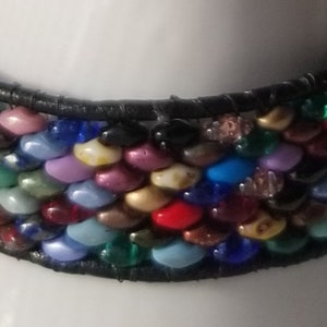 May include: A multi-colored beaded bracelet with a black leather band. The beads are in various colors, including red, blue, green, yellow, and purple. The bracelet is wrapped around a white cylindrical object.