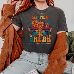 May include: A dark grey t-shirt with a retro-style graphic of sunglasses with a beach scene inside, flowers, and the text "Summer Break #180daysdone #vacaymode".