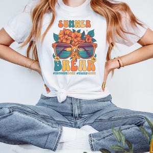 May include: White t-shirt with a retro graphic design featuring sunglasses with a beach scene inside, flowers, and the text "Summer Break #180daysdone #vacaymode".