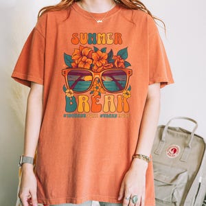 May include: A burnt orange t-shirt with a graphic of sunglasses with a beach scene reflected in the lenses. The sunglasses are surrounded by orange flowers and the text "SUMMER BREAK #100DAYSDONE #VACATIONMODE" is printed below.