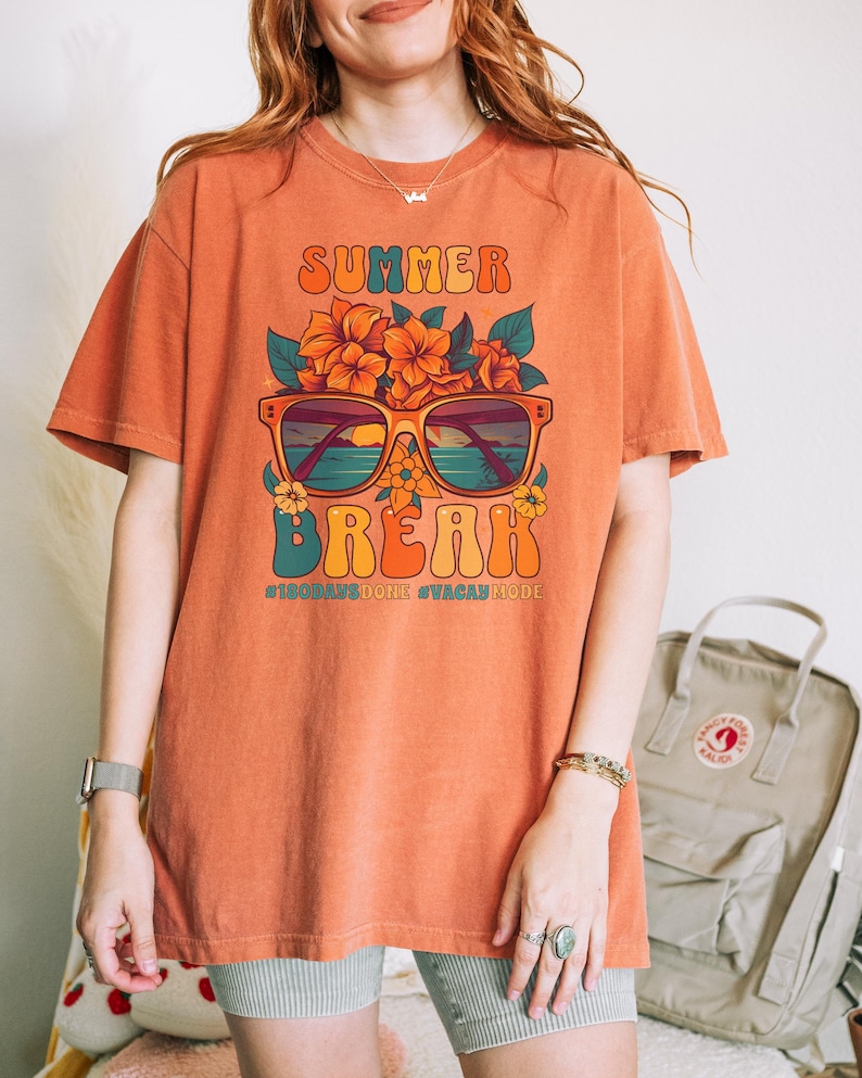 May include: A burnt orange t-shirt with a graphic of sunglasses with a beach scene reflected in the lenses. The sunglasses are surrounded by orange flowers and the text "SUMMER BREAK #100DAYSDONE #VACATIONMODE" is printed below.