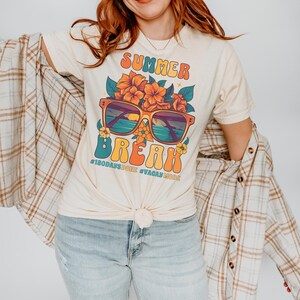 May include: A light beige t-shirt with a retro-style graphic design featuring sunglasses, flowers, and the text "Summer Break #180daysdone #vacaymode".