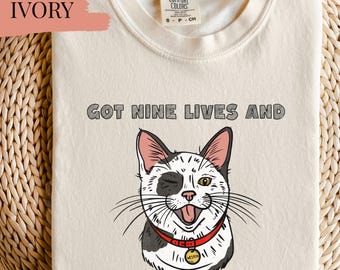 May include: Ivory-coloured t-shirt with a cartoon cat winking and wearing a red collar. The text reads "GOT NINE LIVES AND THEY'RE ALL WEIRD". The shirt is made of a soft material. The t-shirt is a light cream colour.