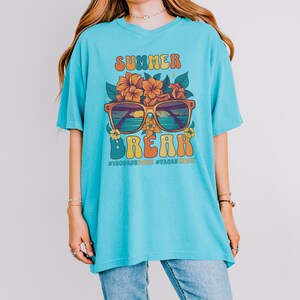 May include: A turquoise blue t-shirt with a retro graphic design featuring the words "Summer Break" in a colorful font. The graphic also includes sunglasses with a beach scene reflected in the lenses and flowers. The text "#180daysdone #vacaymode" is printed below the graphic.