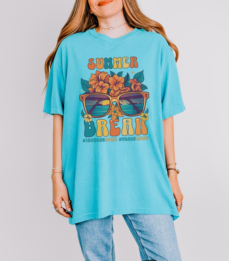 May include: A turquoise blue t-shirt with a retro graphic design featuring the words "Summer Break" in a colorful font. The graphic also includes sunglasses with a beach scene reflected in the lenses and flowers. The text "#180daysdone #vacaymode" is printed below the graphic.