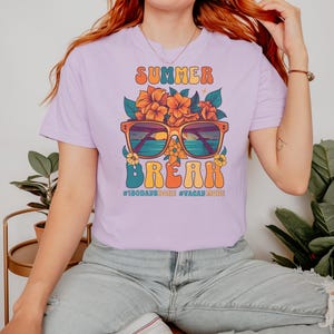 May include: A lavender t-shirt with a graphic design of sunglasses with a sunset and ocean scene, flowers, and the text "Summer Break #180DaysDone #VacayMode".
