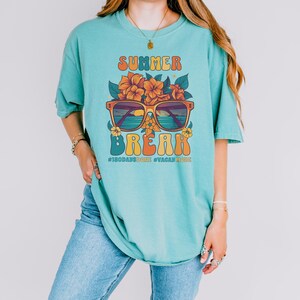 May include: A turquoise blue t-shirt with a retro graphic design featuring sunglasses with a sunset scene, flowers, and the text "Summer Break #180daysdone #vacaymode".