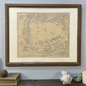 A hand-drawn map of Alagaesia with a white border hangs in a brown wooden frame on a light blue wall. The frame hangs above a wooden shelf that has two books, a pine cone, a feather, two small ceramic birds, and a candle.