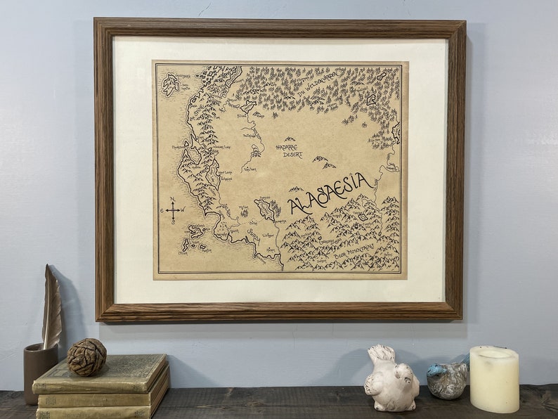A hand-drawn map of Alagaesia with a white border hangs in a brown wooden frame on a light blue wall. The frame hangs above a wooden shelf that has two books, a pine cone, a feather, two small ceramic birds, and a candle.