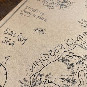 May include: A tan-colored paper map featuring black ink hand-drawn illustrations and text. The map displays the Salish Sea, Whidbey Island, Strait of Juan de Fuca, Smith Island, Minor Island, Deception Pass State Park, and Oak Harbor with a compass rose.