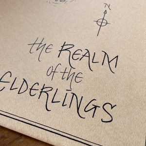 May include: Close-up of a parchment-like paper with the words "The Realm of the Elderlings" in elegant black script. A compass rose and other handwritten details are visible. The paper has a decorative black border.