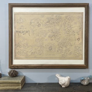 A hand-drawn map of Malazan with a white border hangs in a brown wooden frame on a light blue wall. The frame hangs above a wooden shelf that has two books, a pine cone, a feather, two small ceramic birds, and a candle.