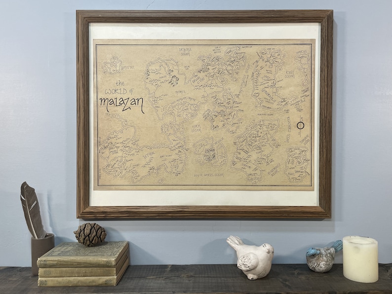 A hand-drawn map of Malazan with a white border hangs in a brown wooden frame on a light blue wall. The frame hangs above a wooden shelf that has two books, a pine cone, a feather, two small ceramic birds, and a candle.