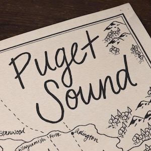 May include: A cream-colored paper map featuring black ink illustrations of Puget Sound. The map displays hand-lettered text including Puget Sound, Skagit River, Stanwood, Stillaguamish River, and Arlington, alongside drawings of mountains and trees.