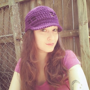 May include: A woman with long brown hair is wearing a purple crocheted hat with a brim. The hat has a crocheted design with open spaces. She is wearing a pink shirt.