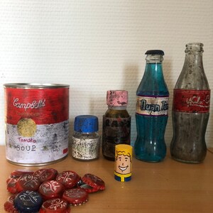 Peut inclure: Une collection d'objets sur le thème de Fallout, dont une boîte de soupe tomate Campbell's, une bouteille de Nuka Cola, une bouteille de Sunset Sarsaparilla, une bouteille de Rad-Away, une bouteille d'eau purifiée et une figurine de Vault Boy. Il y a aussi plusieurs capsules de bouteilles avec le logo Nuka Cola.
