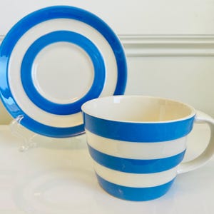 May include: A ceramic cup and matching saucer set featuring a bold pattern of horizontal blue and white stripes. The cup has a wide, rounded body with a single handle, while the saucer displays concentric blue and white circular bands.