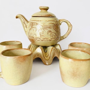 May include: A ceramic tea set featuring a teapot with a lid and four matching mugs. The pieces are light tan with darker brown accents and intricate embossed geometric patterns on the teapot. The teapot rests on a wavy, sculpted base.