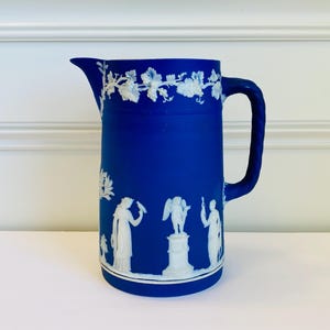 May include: A cobalt blue pitcher with a white relief design. The pitcher features a handle and spout, with a decorative border of grapevines. The body of the pitcher showcases classical figures and a pedestal statue, creating a vintage aesthetic.