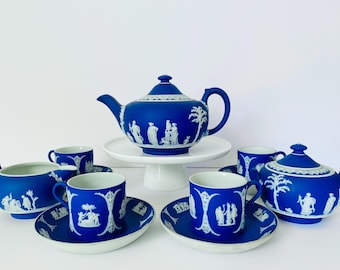 May include: A blue and white tea set with a teapot, creamer, sugar bowl, and four cups and saucers. The set features a white cameo design of figures and trees on a cobalt blue background.