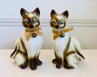 May include: Two ceramic Siamese cat figurines. The cats are seated with a white body, brown markings, and a gold ribbon and bell around their necks. The figurines have blue eyes and are on a white surface.