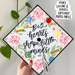 Puede incluir: Un gorro de graduación decorado con un patrón floral e iconos con temática escolar. El gorro tiene el texto "Big hearts shape little minds" escrito en él. El año 2023 también está escrito en el gorro.