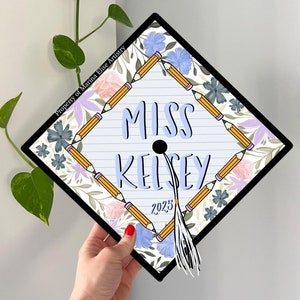 Puede incluir: Un gorro de graduación azul y blanco con un diseño floral y lápices. El gorro dice "Miss Kelsey" y "2023".