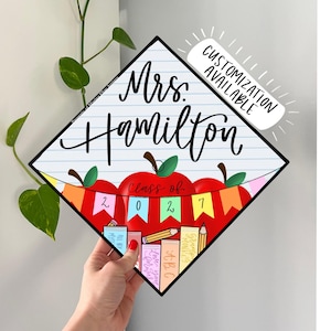 Könnte beinhalten: Ein rautenförmiger Abschlusskappen-Aufsatz mit weißem linierten Papierhintergrund und dem Text Mrs. Hamilton in schwarzer Schreibschrift. Darunter zeigt eine rote Apfelgrafik ein buntes Banner mit dem Text Class of 2027 und gestapelten Büchern.