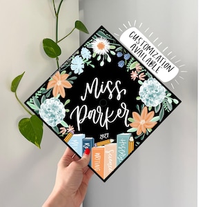 May include: A black graduation cap topper featuring a floral wreath with peach and blue flowers, the text "Miss Parker" in white script, the year "2027", and a stack of books labeled "Math", "Writing", "Science", and "History" with a pencil.